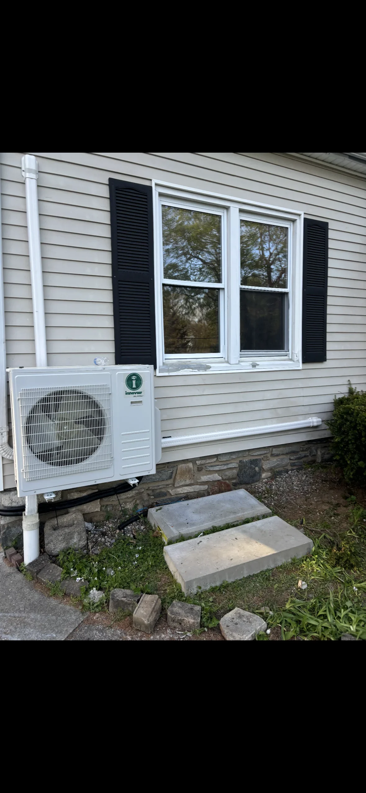White mini split heat pump condenser unit installed below residential windows with black shutters